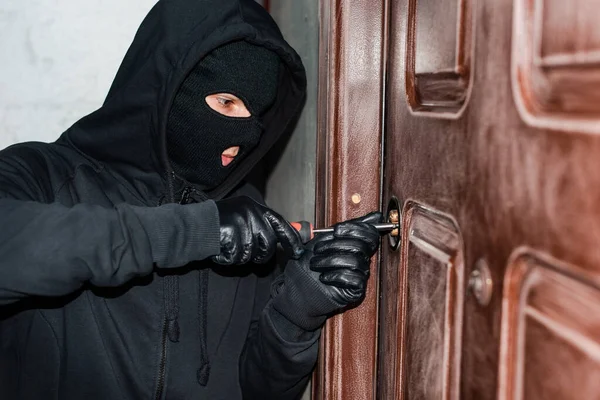 Burglar in balaclava and leather gloves using screwdriver while breaking door lock