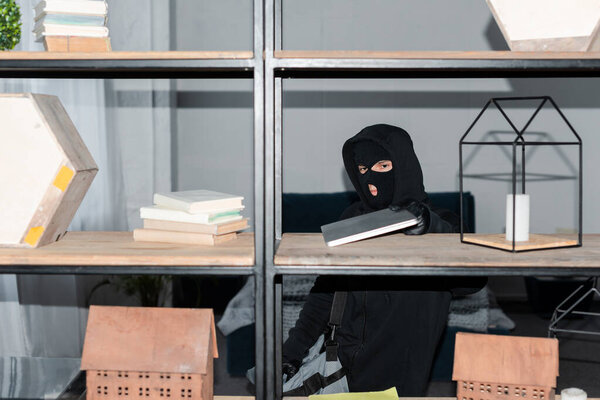 Thief in mask and leather gloves taking laptop from cupboard during stealing 