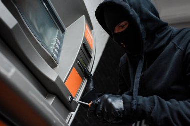 Low angle view of thief in balaclava using screwdriver while breaking atm  clipart