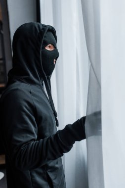 Perdelerin yanında duran kar maskeli soyguncunun yan görüntüsü 