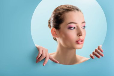 Parlak dudakları olan güzel çıplak bir kadının portresi, pembe göz farı, mavi üzerine izole edilmiş kağıttan yapılmış bir delikten başka tarafa bakıyor.