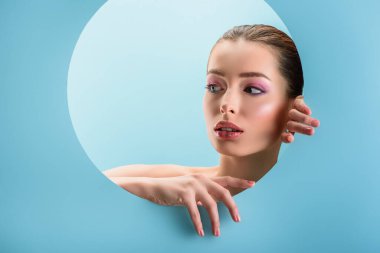portrait of beautiful naked woman with glossy lips, pink eyeshadow looking through paper round hole isolated on blue clipart