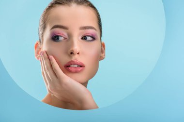 Parlak dudakları olan güzel bir genç kadının portresi, pembe göz farı, mavi üzerine izole edilmiş kağıttan yapılmış bir delikten başka tarafa bakıyor.