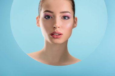Parlak dudakları olan güzel çıplak bir kadının portresi, pembe göz farı, mavi üzerine izole edilmiş kağıttan yuvarlak bir delikten bakıyor.