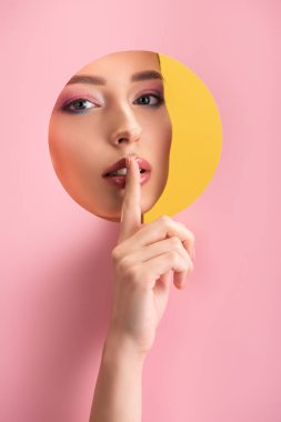 portrait of beautiful woman with shiny makeup in pink paper round hole showing shh sign isolated on yellow