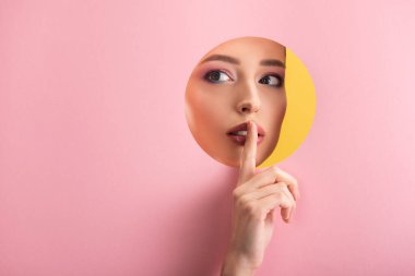 portrait of beautiful woman with shiny makeup in pink paper round hole showing shh sign isolated on yellow clipart