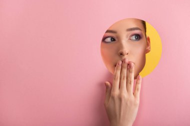 portrait of beautiful woman with shiny makeup in pink paper round hole covering mouth with hand isolated on yellow clipart