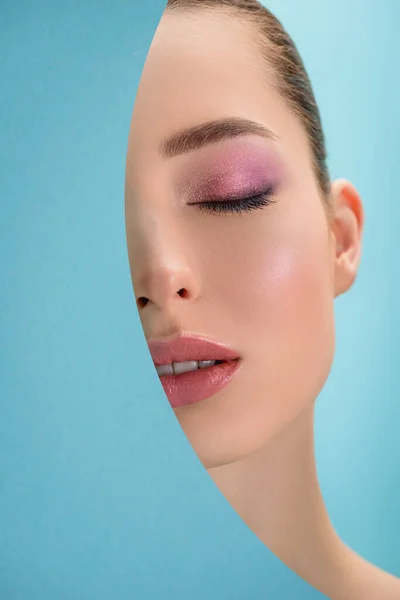 portrait of beautiful woman with closed eyes, glossy lips, pink eyeshadow behind paper round hole isolated on blue