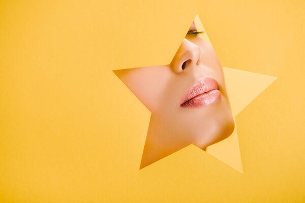cropped view of beautiful woman with shiny lips in paper star shaped hole isolated on yellow
