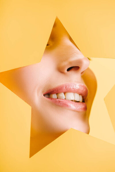 cropped view of beautiful woman with shiny lips in paper star shaped hole smiling isolated on yellow