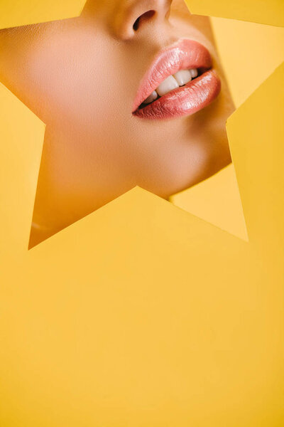 cropped view of beautiful woman with shiny lips in paper star shaped hole isolated on yellow