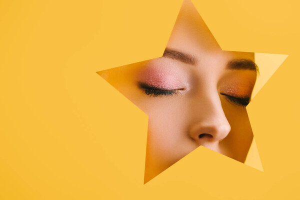 portrait of beautiful woman with shiny makeup in paper star shaped hole with closed eyes isolated on yellow