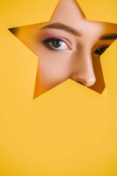 portrait of beautiful woman with shiny makeup in paper star shaped hole looking at camera isolated on yellow