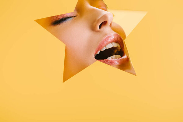 portrait of beautiful woman with shiny makeup in paper star shaped hole with closed eyes and open mouth isolated on yellow