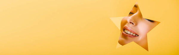 portrait of beautiful woman with shiny makeup in paper star shaped hole smiling isolated on yellow, panoramic shot