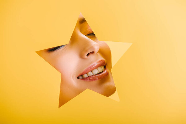 portrait of beautiful woman with shiny makeup in paper star shaped hole smiling isolated on yellow