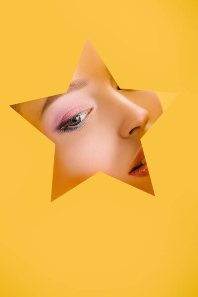 portrait of beautiful woman with shiny makeup in paper star shaped hole looking away isolated on yellow