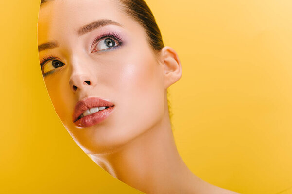 portrait of beautiful woman with shiny makeup in paper round hole looking up isolated on yellow