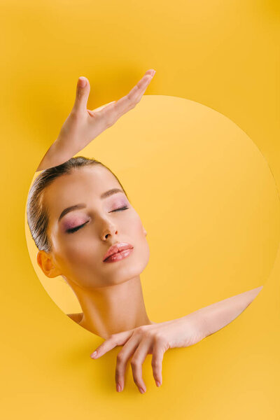 portrait of beautiful woman with shiny makeup in paper round hole with hands and closed eyes isolated on yellow