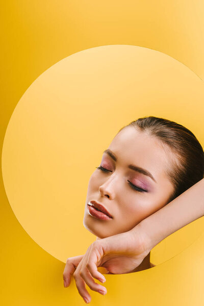 portrait of beautiful woman with shiny makeup in paper round hole with hand and closed eyes isolated on yellow