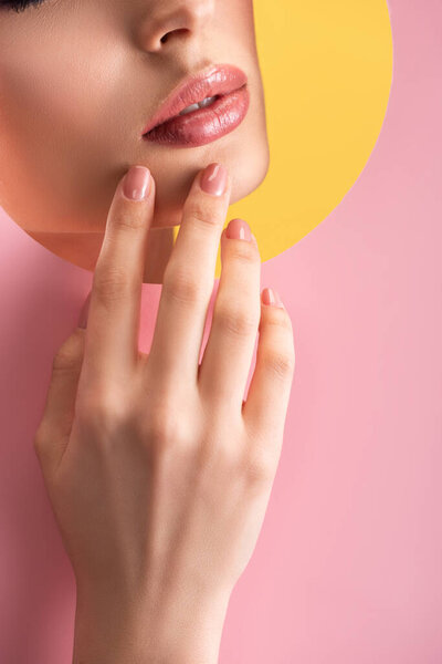 partial view of beautiful woman with shiny lips in pink paper round hole with hand isolated on yellow