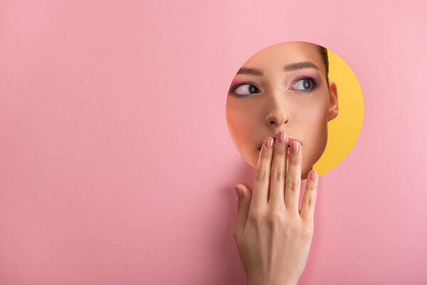 portrait of beautiful woman with shiny makeup in pink paper round hole covering mouth with hand isolated on yellow