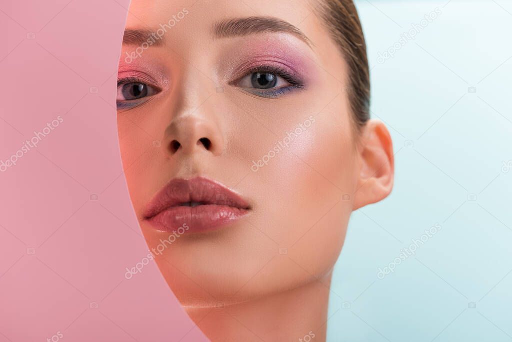 portrait of beautiful woman with shiny makeup looking through round paper hole isolated on blue