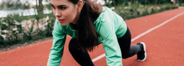 Panoramic crop of attractive sportswoman standing in starting position before running on track in park  clipart