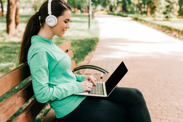 在公园的长椅上使用笔记本电脑的耳机中迷人的微笑自由职业者的侧视图 — 图库照片