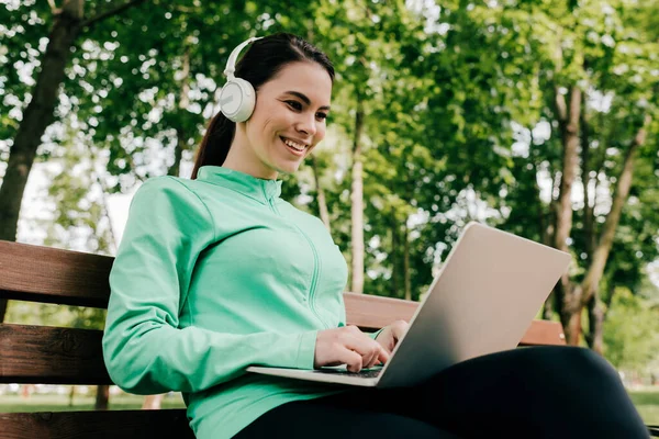 在公园的长椅上用笔记本电脑对着耳机笑的女孩 — 图库照片
