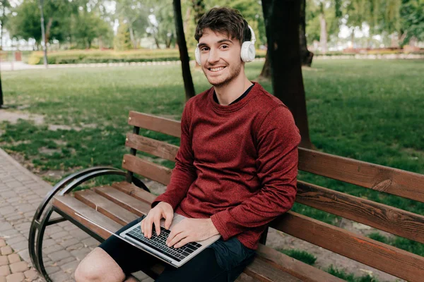 Uomo Sorridente Cuffia Guardando Fotocamera Durante Utilizzo Laptop Nel Parco — Foto Stock