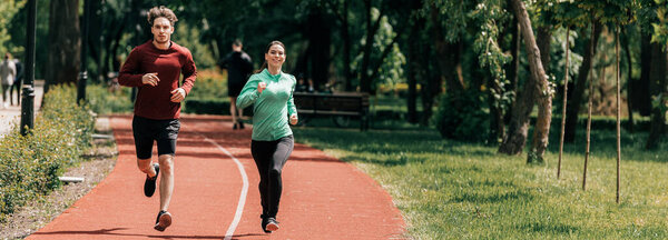 Panoramic shot of smiling woman jogging near boyfriend on running track in park 