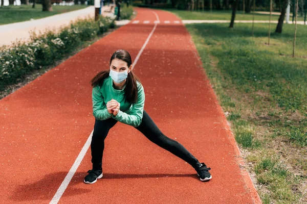 Sportswoman in medical mask exercising on running track in park