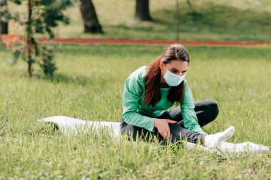 Sağlık maskeli sportmen kadının seçici odağı parktaki spor minderinin üzerinde geriniyor. 