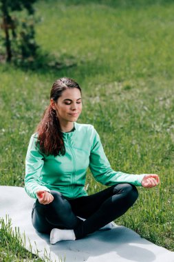 Çekici bir kadın parkta çimlerin üzerinde fitness minderi üzerinde meditasyon yapıyor. 