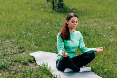 Bacak bacak üstüne atmış güzel bir kadın parkta çimlerin üzerinde meditasyon yapıyor. 