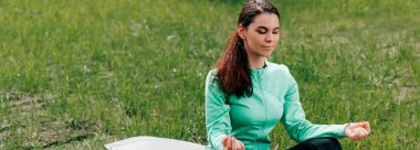 Parktaki spor minderi üzerinde meditasyon yapan panoramik genç bir kadın. 