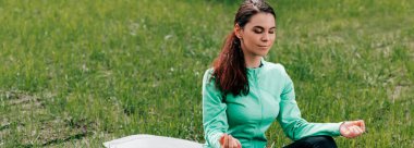 Panoramic crop of young woman meditating on fitness mat in park  clipart
