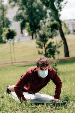 Tıbbi maskeli genç sporcunun seçici odak noktası parktaki çimlerin üzerindeki spor minderinde şınav çekiyor. 