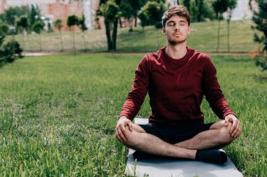 Yakışıklı genç adam parkta fitness minderinde otururken meditasyon yapıyor. 