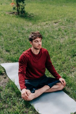 Gözleri kapalı yakışıklı bir adam yoga yaparken parkta spor minderinde poz veriyor. 