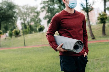 Sağlık maskeli sporcunun parkta spor minderi tutuşu. 