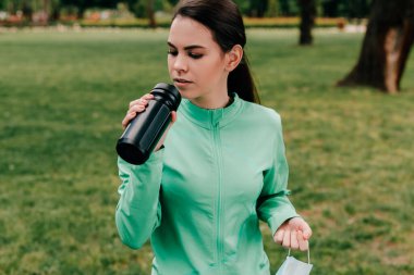 Parkta spor şişesi ve sağlık maskesi tutan çekici bir sporcu kadın. 