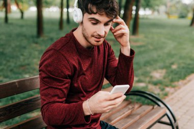 Yakışıklı sporcu kulaklıkla müzik dinliyor ve parkta bankta akıllı telefon kullanıyor. 