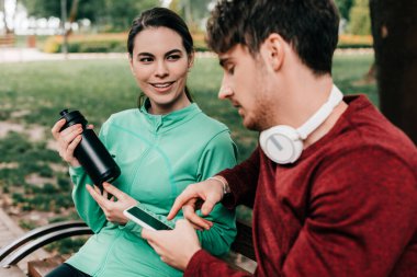 Parktaki bankta akıllı telefon kullanarak spor şişesini erkek arkadaşının yanında tutan gülümseyen sporcu kadının seçici odak noktası. 