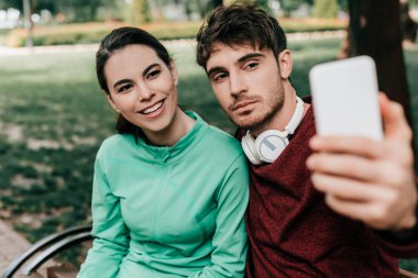 Parktaki bankta oturan yakışıklı erkek arkadaşın yanında akıllı telefonuyla selfie çeken gülümseyen sporcu kadının seçici odağı. 