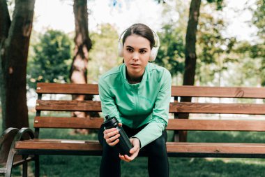 Kulaklıklı çekici bir sporcu. Parktaki bankta spor şişesi tutuyor. 