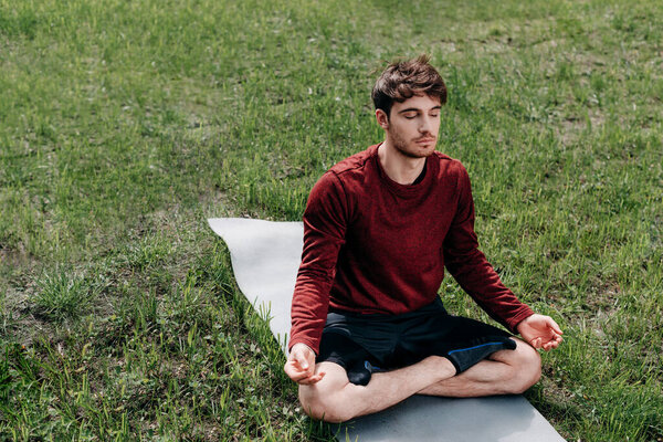 Young man meditating on fitness mat on grass in park 