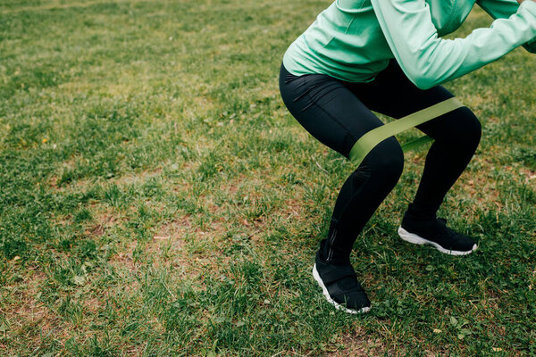 Cropped view of sportswoman doing squat with resistance band on grass in park 
