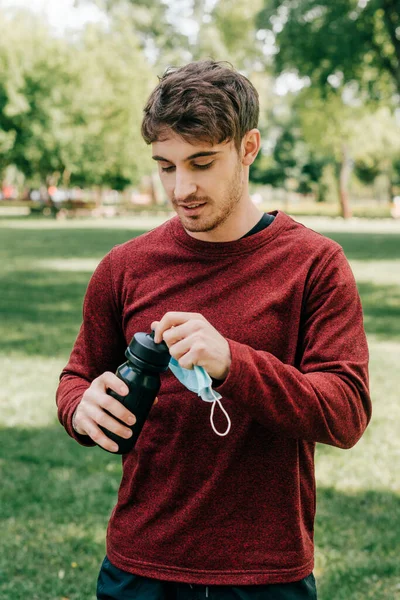 Guapo Deportista Sosteniendo Máscara Médica Abriendo Botella Deportiva Parque — Foto de Stock